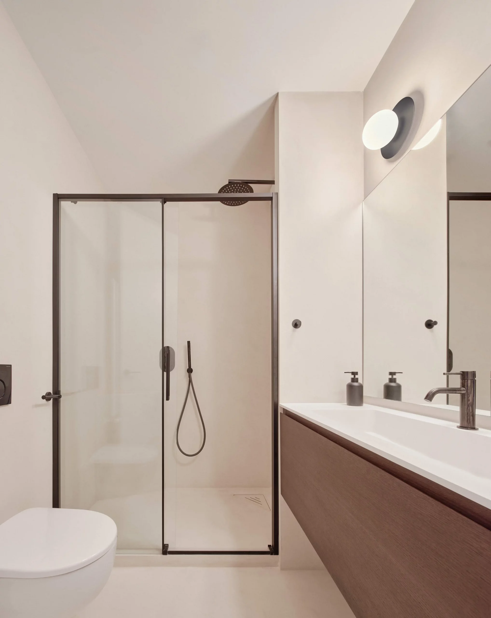 Minimalist bathroom with microcement walls, glass shower, and wooden vanity by Cometa Architects