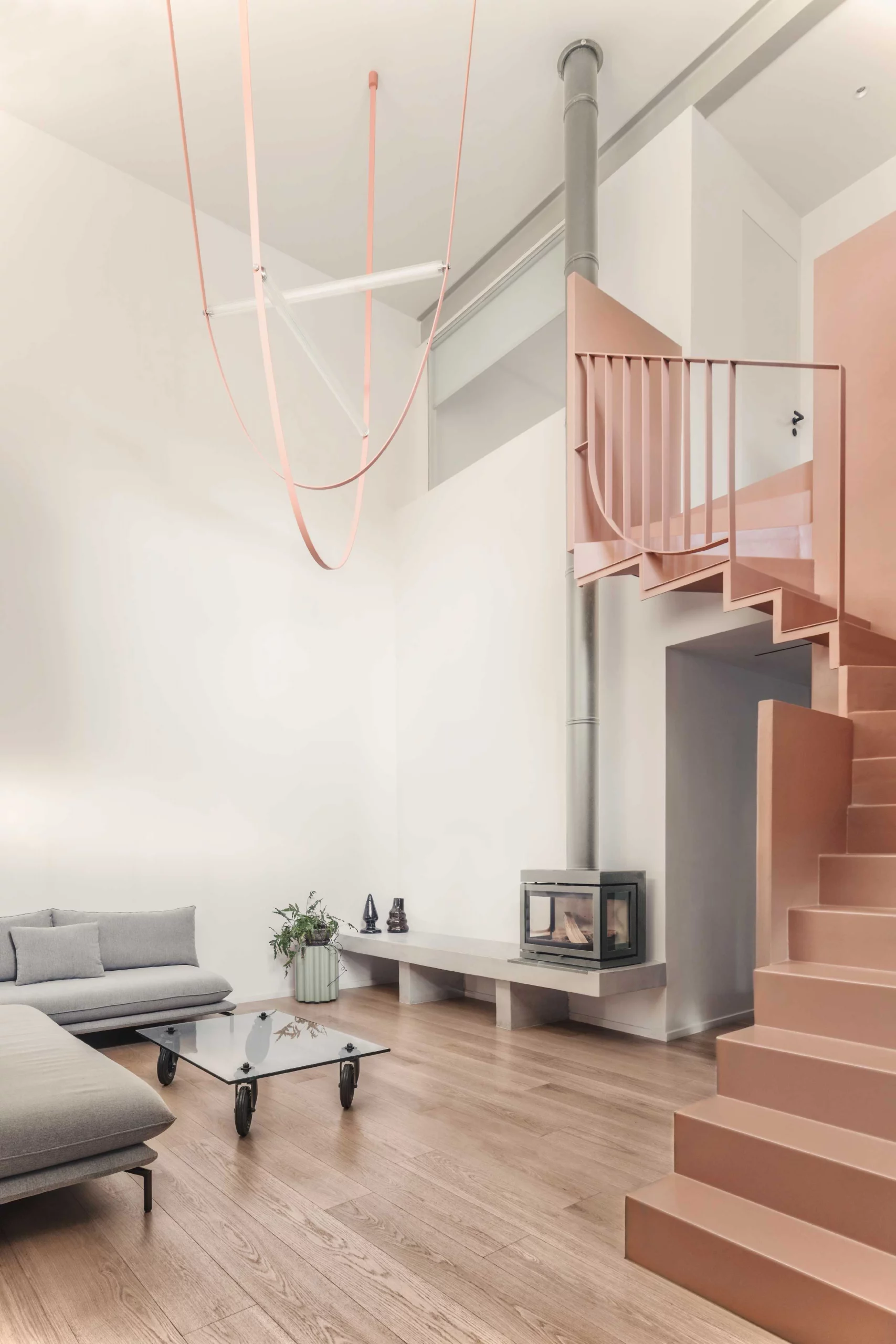 Double-height living space of Casa D by Cometa Architects in Athens featuring a pink steel staircase, minimalist lighting, grey sofa, and wood flooring.