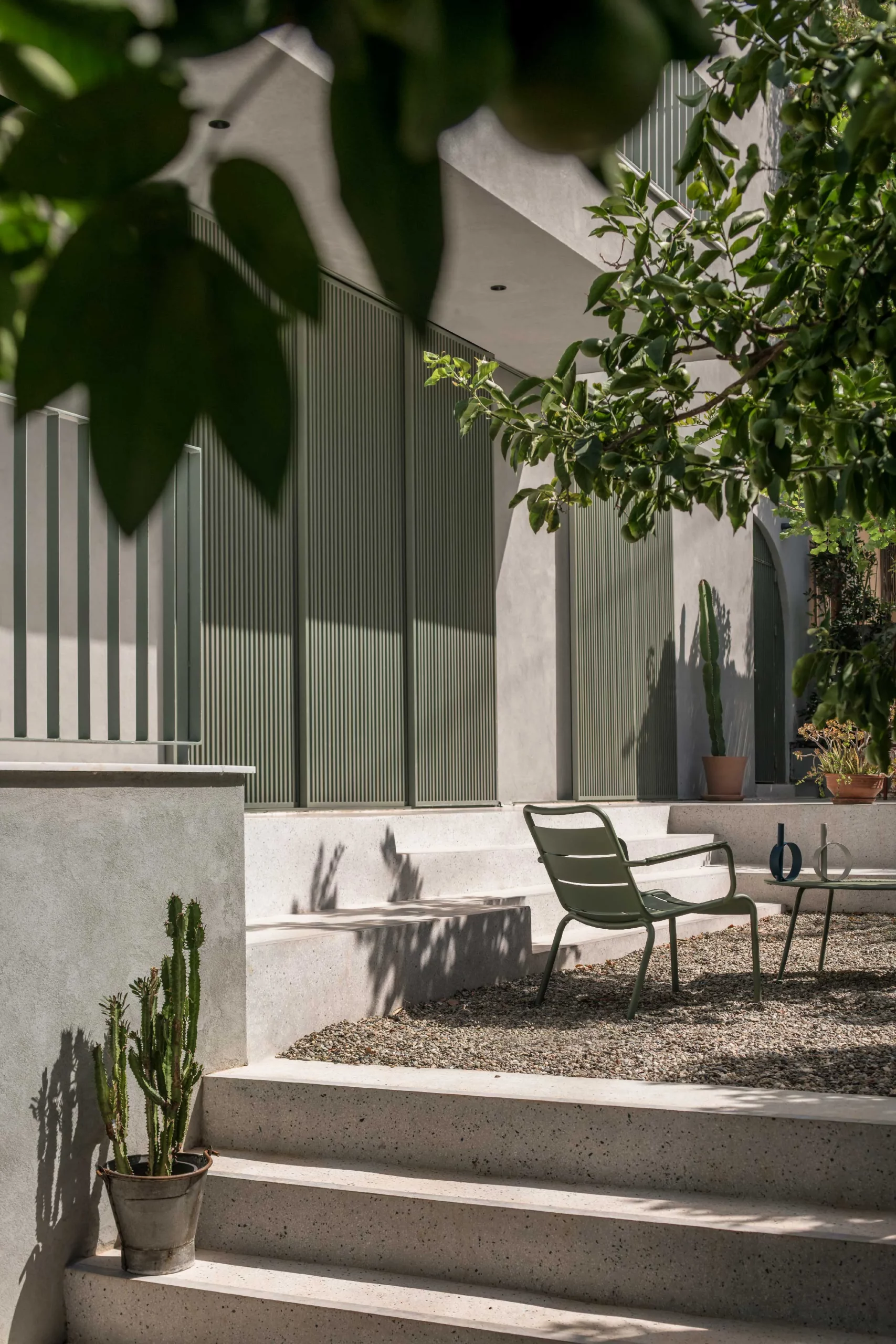 Outdoor dining area of Casa D by Cometa Architects in Athens with red slatted table, green aluminum pergola, and arched door casting dappled shadows on plastered wall.