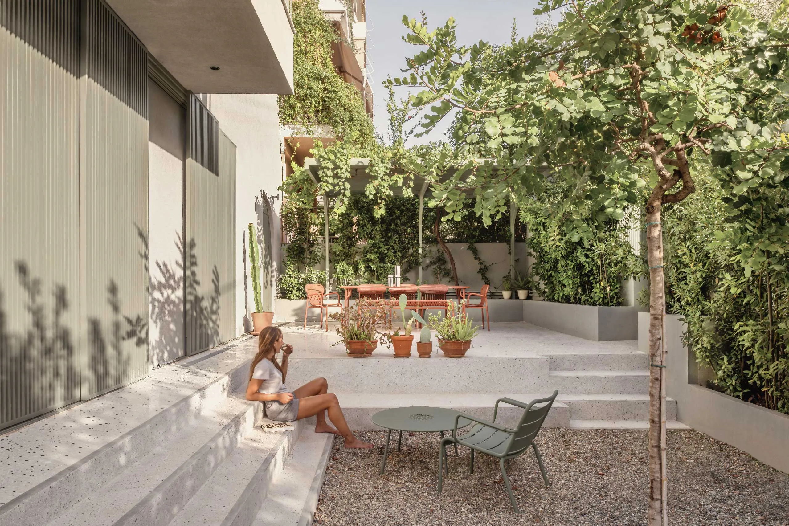 Detail of Casa D garden by Cometa Architects in Athens showing terrazzo steps, green aluminum shutters, cactus in terracotta pots, and outdoor chair under Mediterranean light.