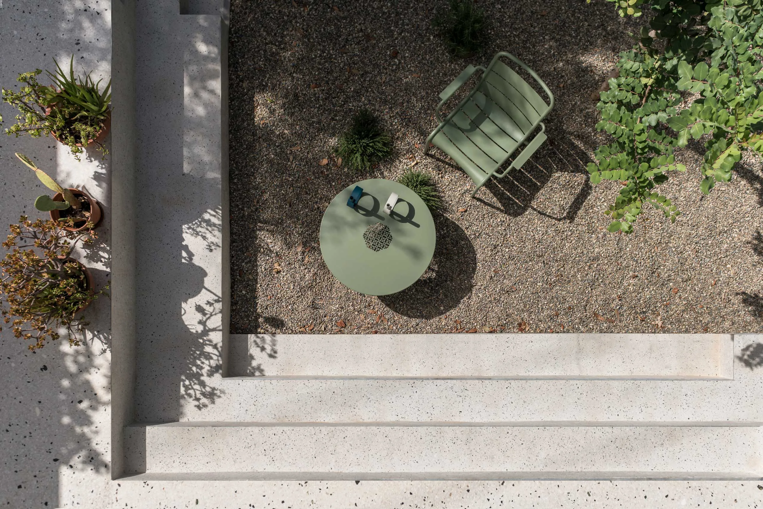 Aerial view of Casa D garden by Cometa Architects in Athens showing terrazzo steps, green metal chair and table, and terracotta pots with Mediterranean plants.