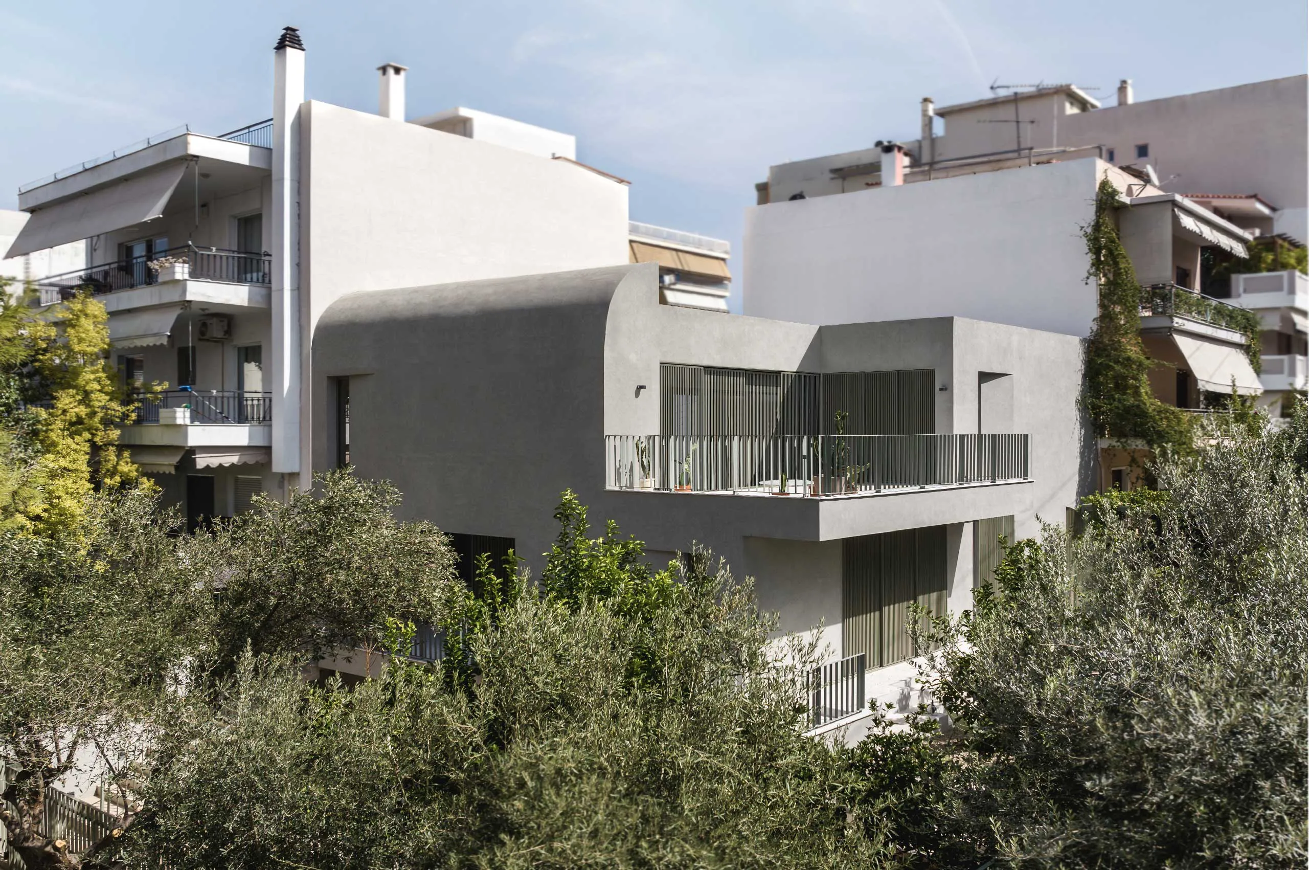 Exterior view of Casa D by Cometa Architects in Athens showing curved grey plastered volume surrounded by urban buildings and Mediterranean vegetation.