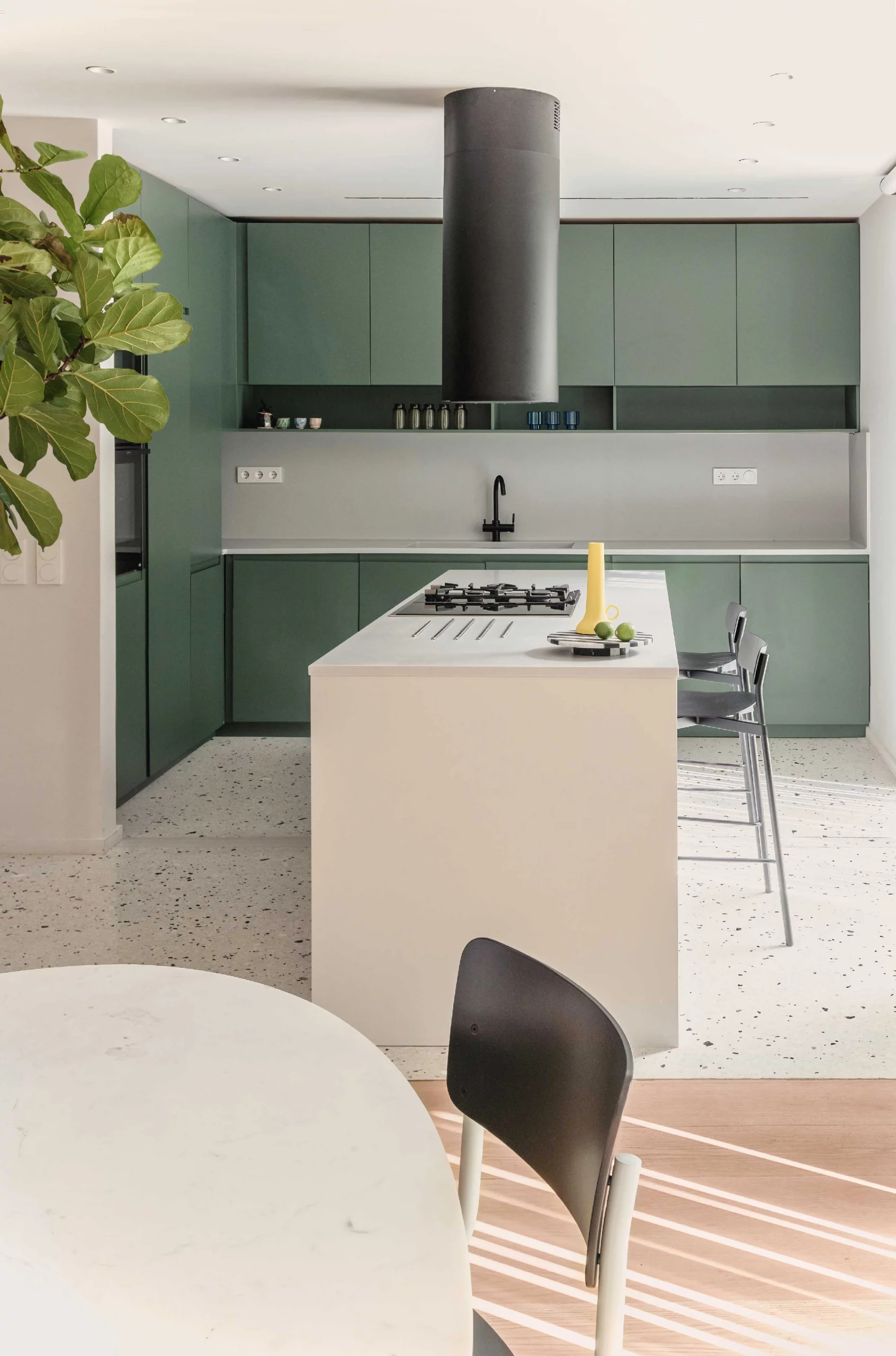 Kitchen and dining area of Casa D by Cometa Architects in Athens featuring green cabinetry, white island, terrazzo floor, and black cylindrical extractor.