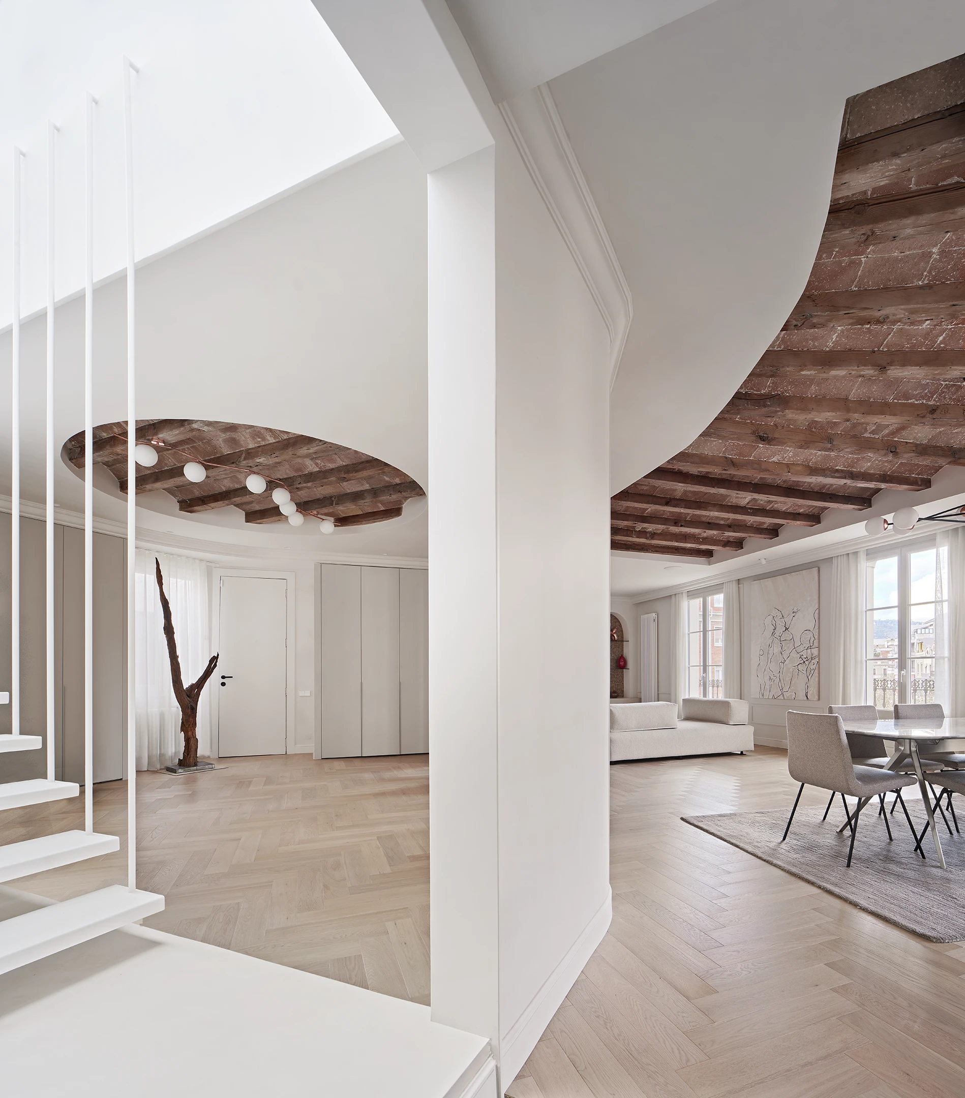 Open-plan living area with dining space, floating staircase, and exposed wooden beams and terracotta tiles in a Barcelona apartment.