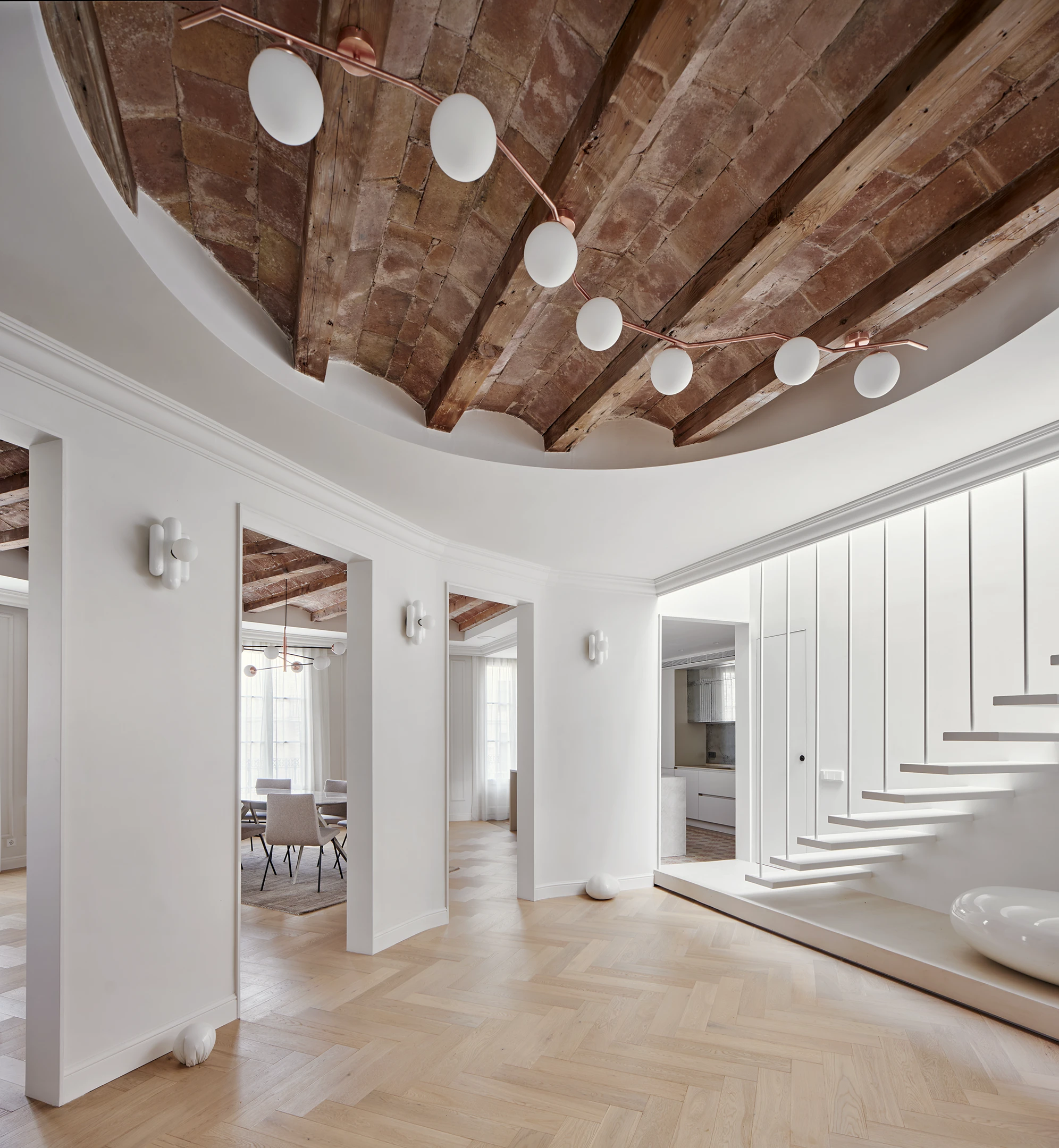 Entrance hall with floating staircase, circular ceiling cut-out, and original wooden beams and terracotta tiles in a Barcelona apartment.