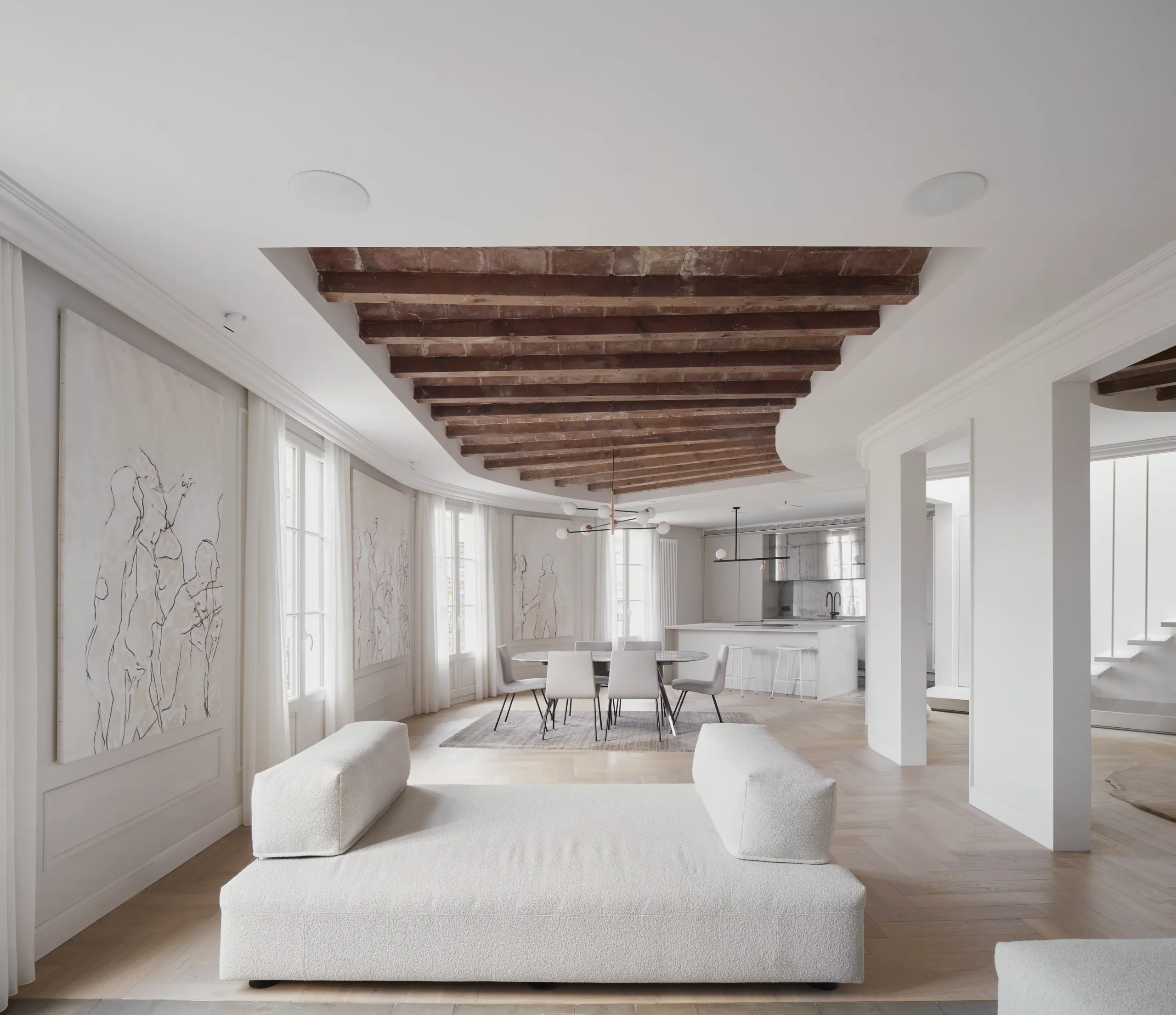 Open-plan living space with curved layout, wooden beams and terracotta tiles, minimalist furniture, and a view of the kitchen in a Barcelona apartment.