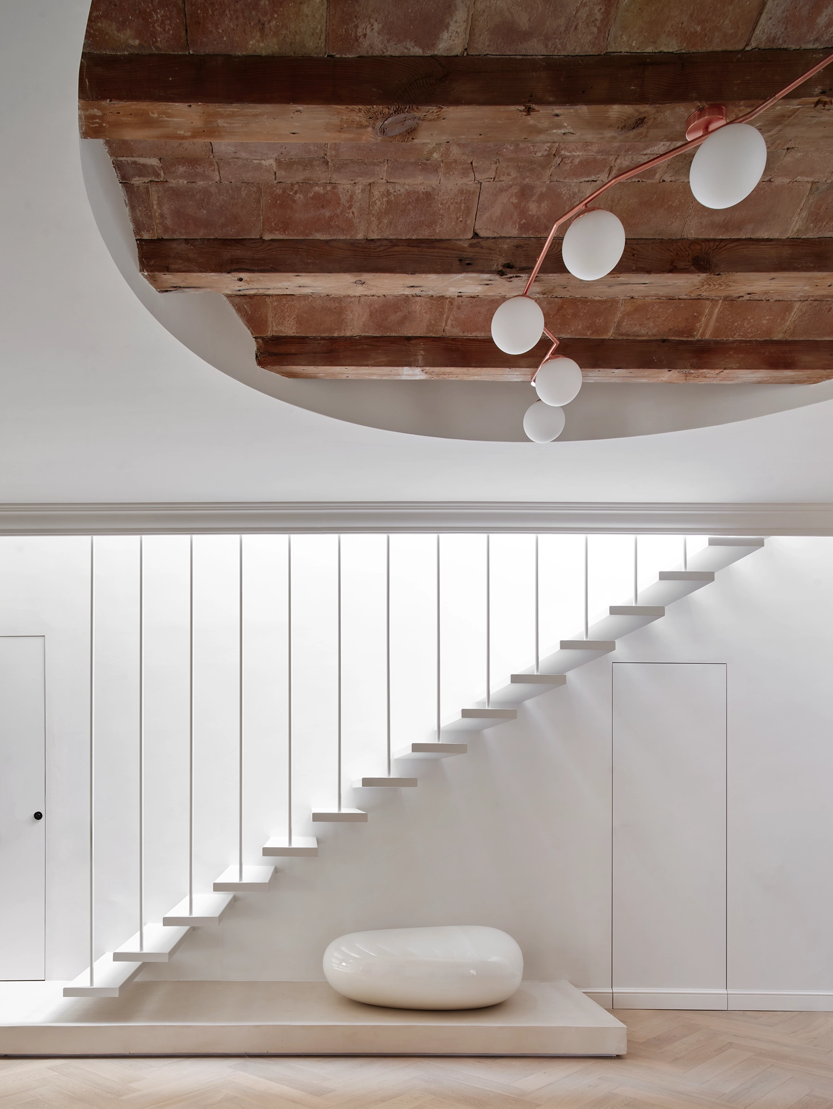 White floating staircase with vertical balustrades, circular ceiling cut-out, and wooden beams and terracotta tiles in a Barcelona apartment.
