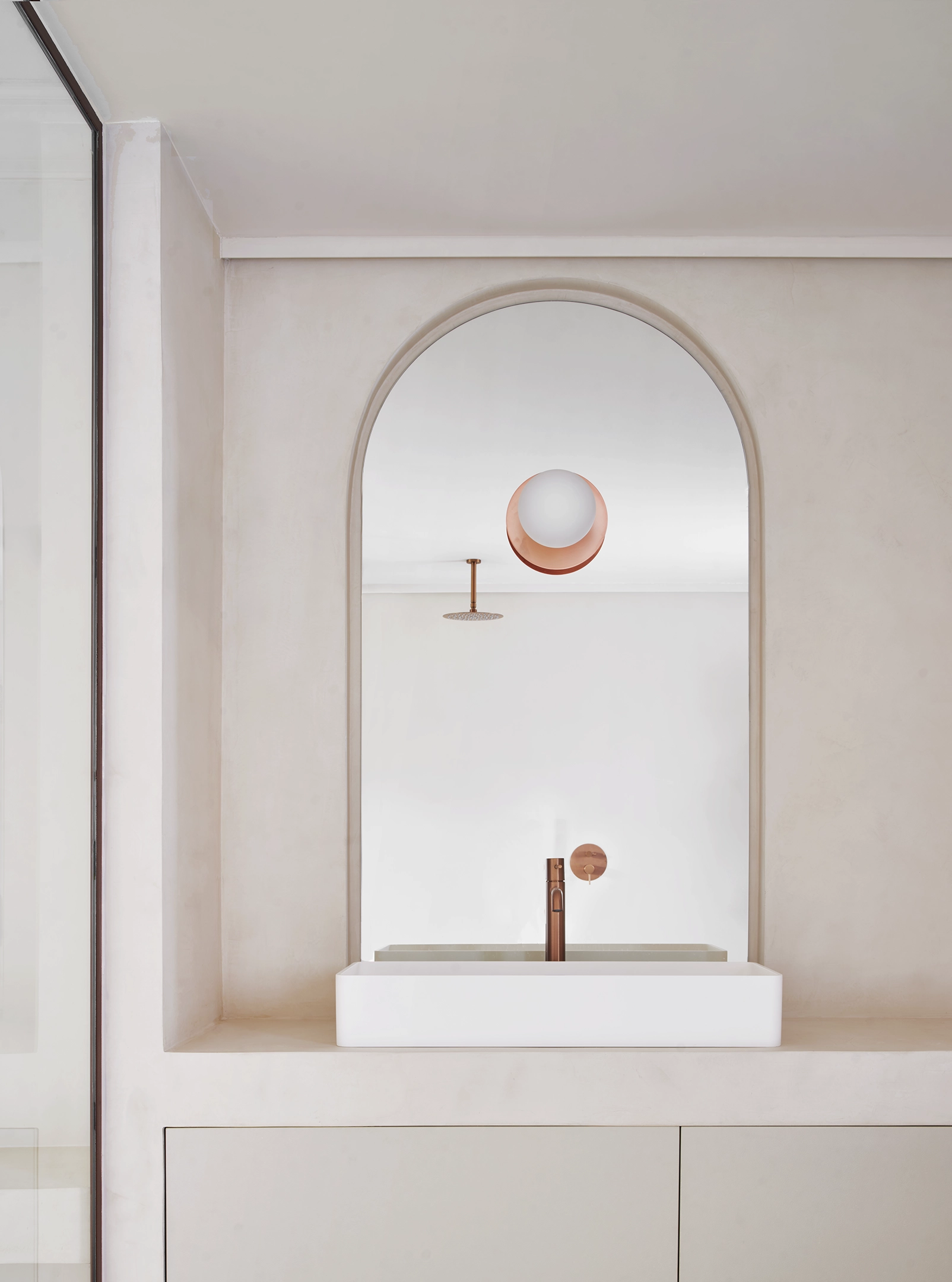 Arched mirror and white sink with copper faucet in a minimalist wetroom bathroom in a Barcelona apartment.