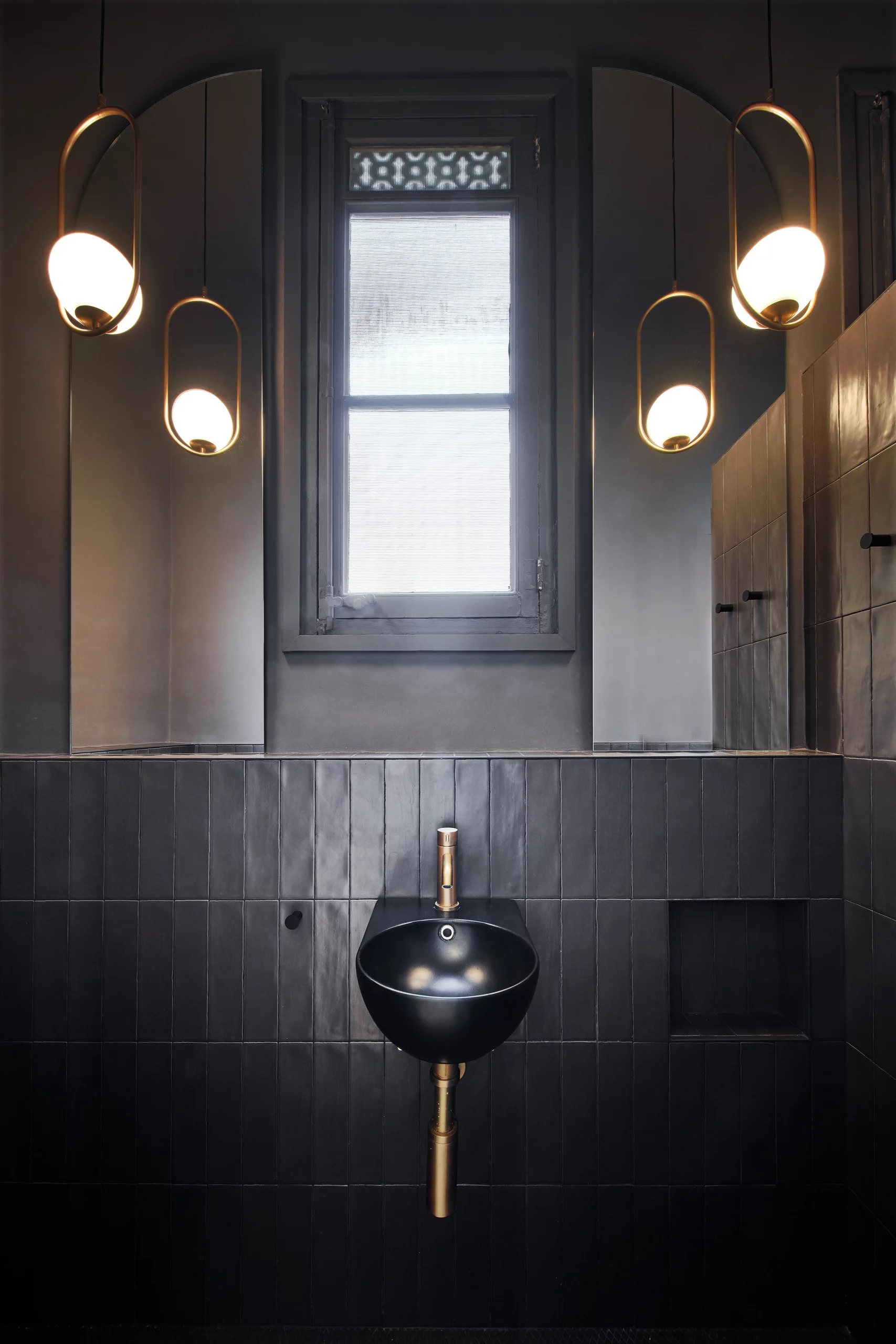 Black ceramic sink with brass fixtures and pendant lights in a dark-tiled bathroom by Cometa Architects in Barcelona.