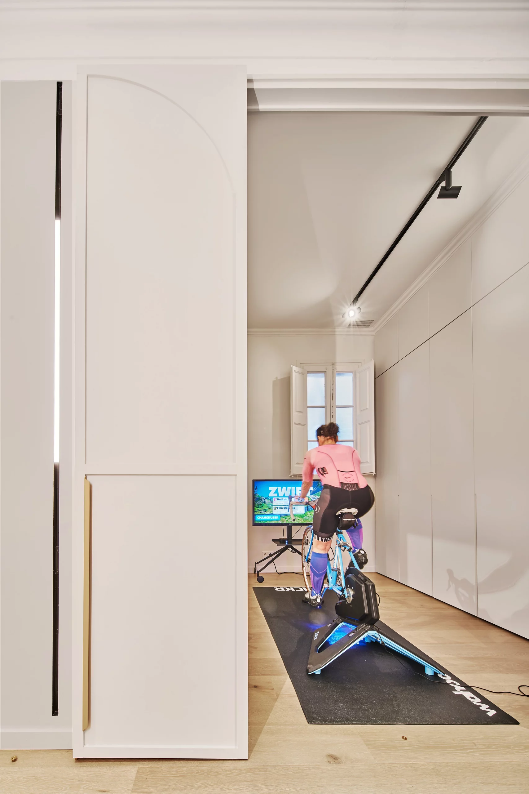 Woman training on a bike inside a minimalist white home gym with custom sliding door by Cometa Architects in Barcelona.