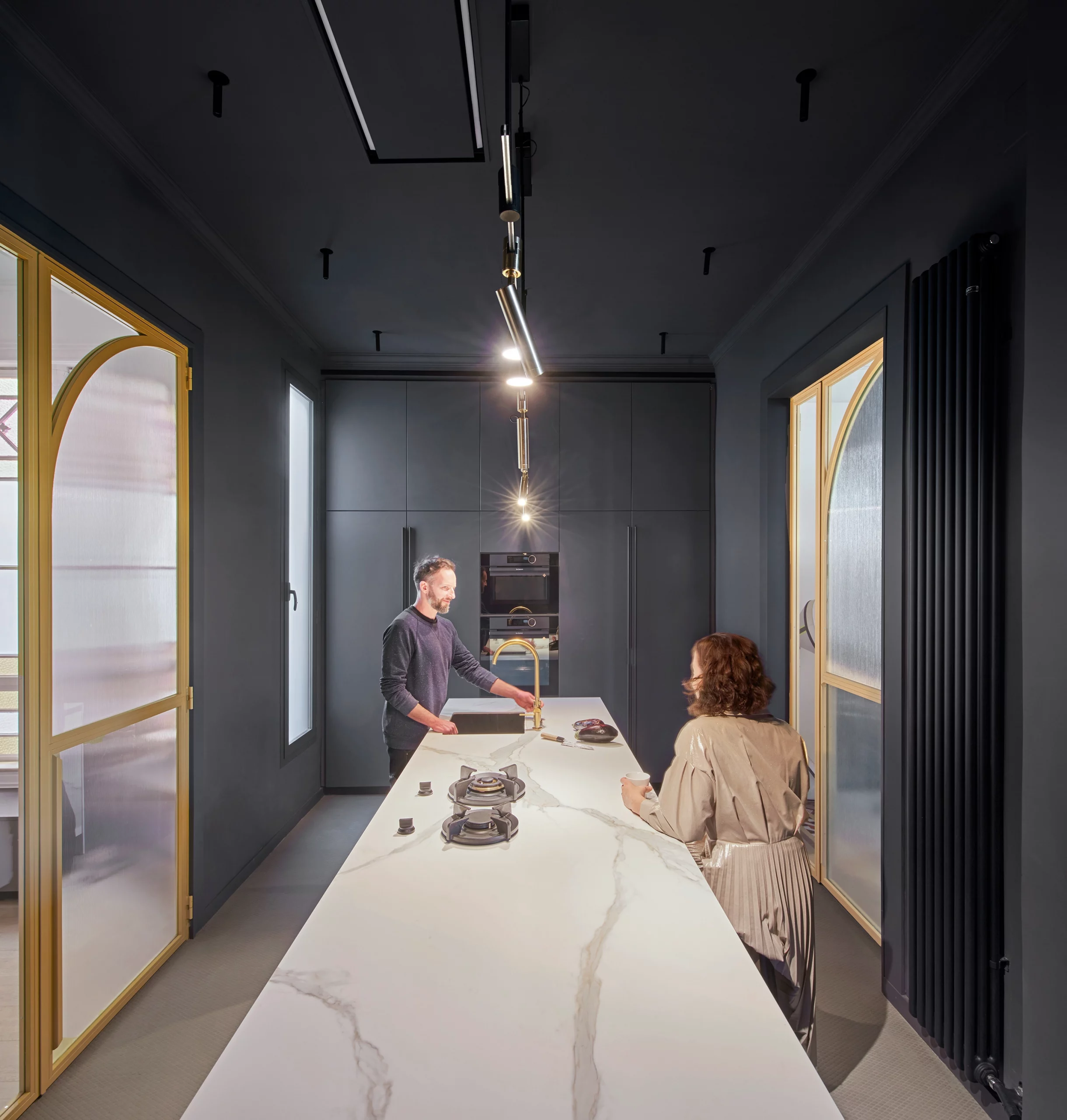 Couple interacting around a white marble kitchen island with brass faucet and dark cabinets by Cometa Architects in Barcelona.