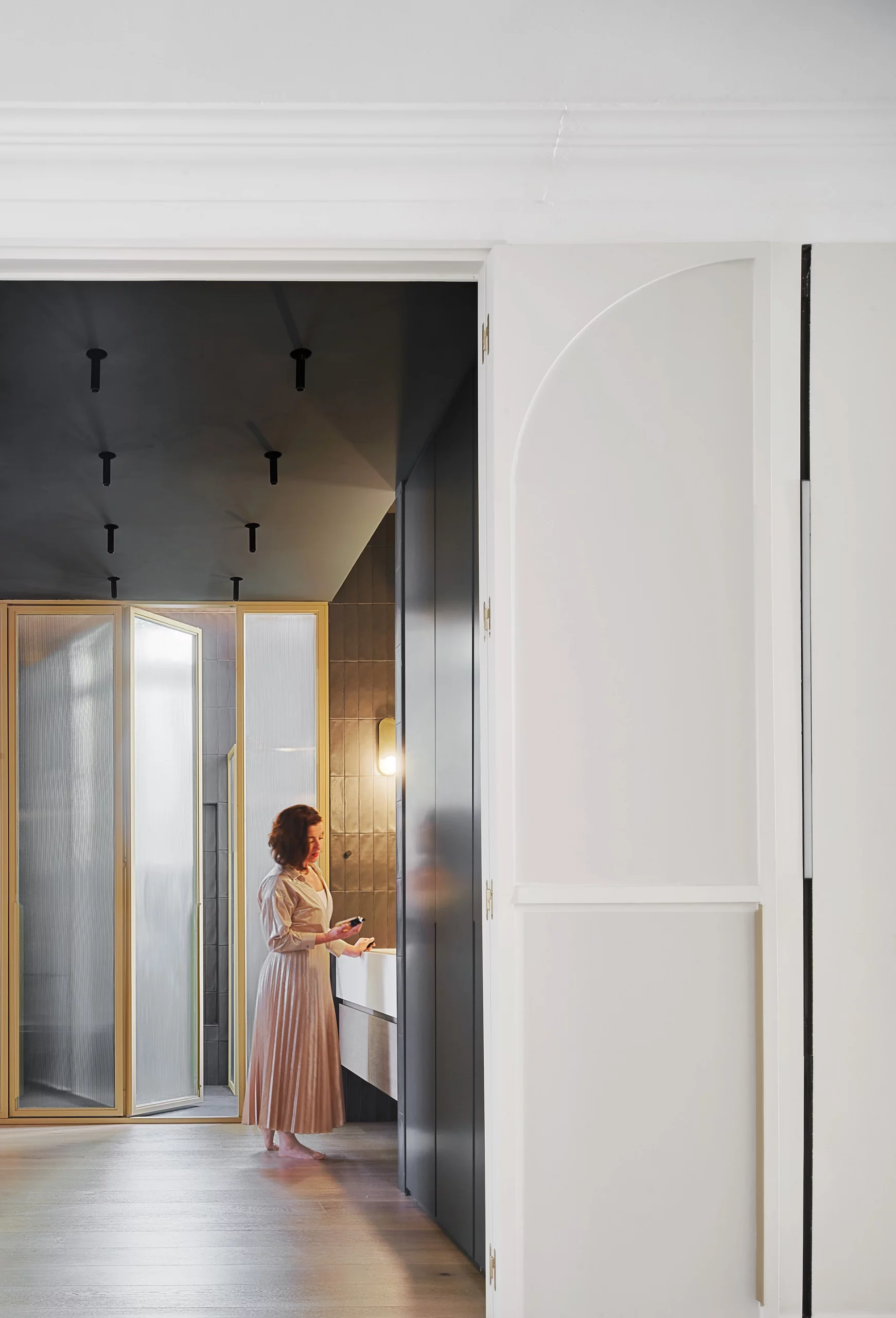 Woman standing at the vanity through an arched sliding door connecting the bright corridor and the dark-toned bathroom, designed by Cometa Architects.