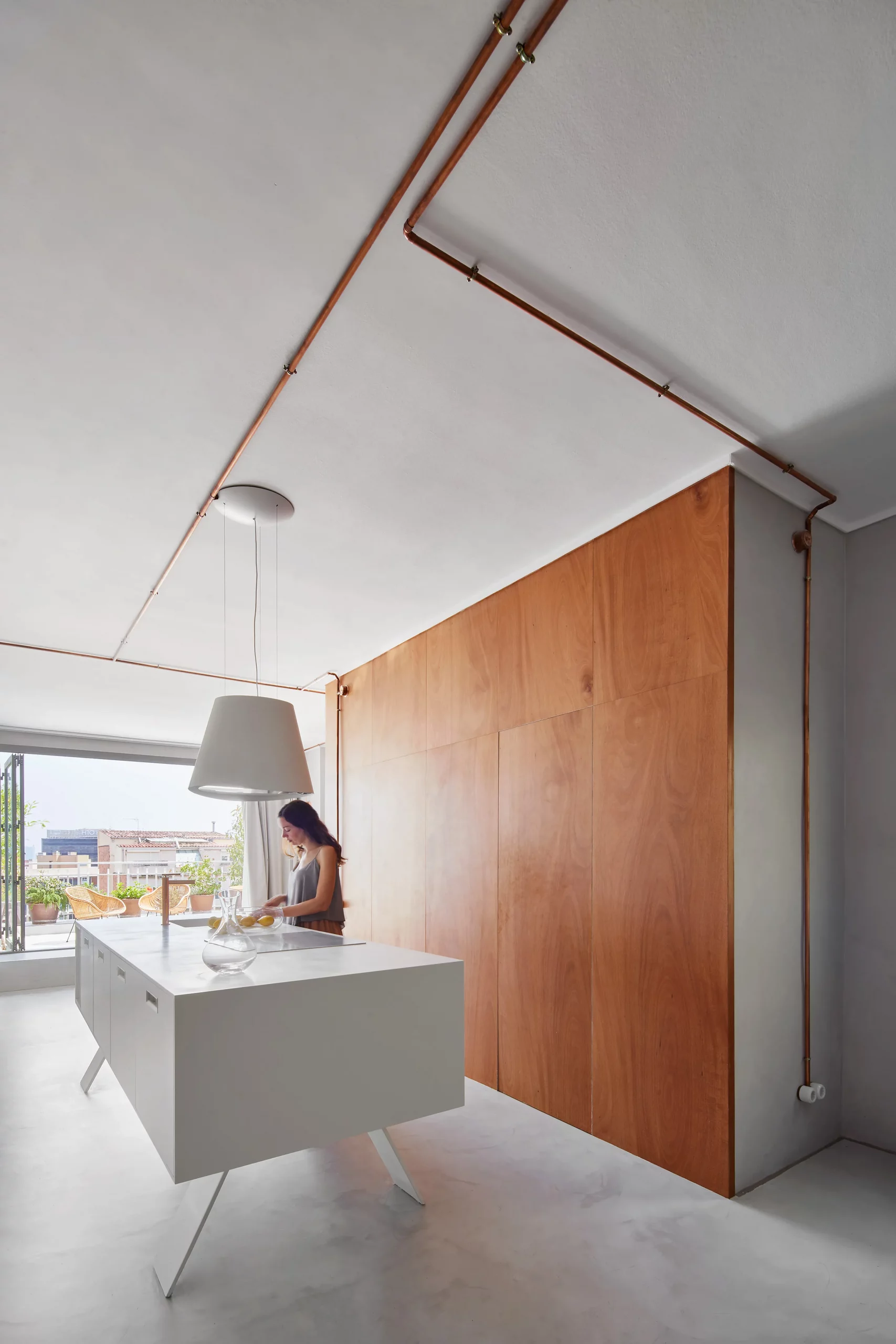 Minimalist kitchen with white island, copper piping, and wooden wall by Cometa Architects in Barcelona