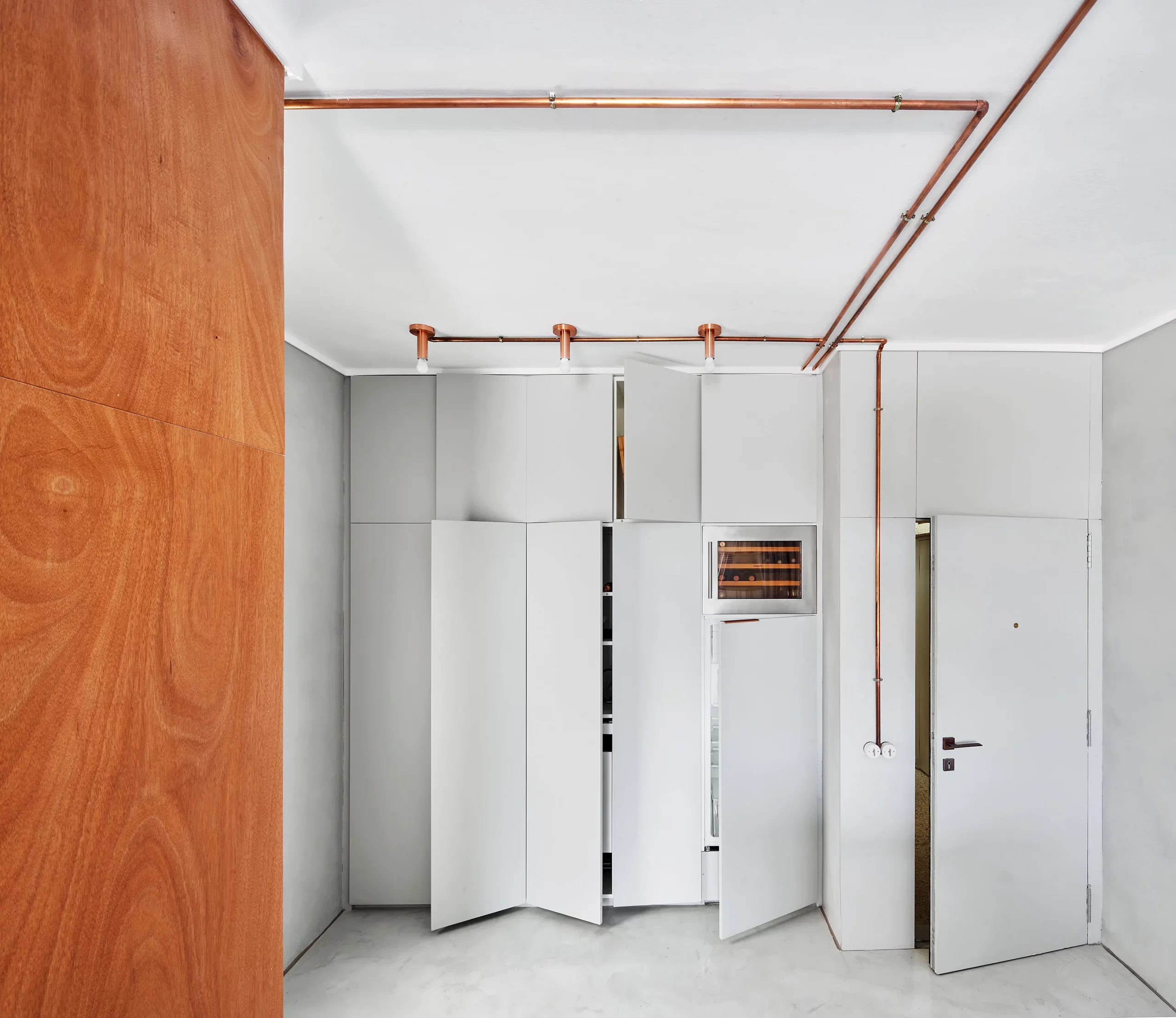 Custom white kitchen cabinets with copper piping and wooden wall detail by Cometa Architects in Barcelona