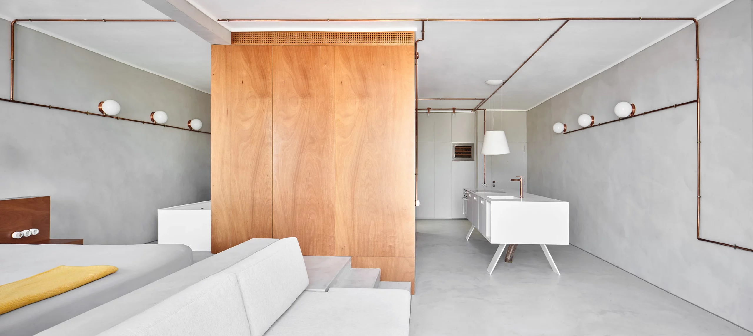 Interior view of Marina Apartment showing bedroom, bathtub, and kitchen island with copper piping by Cometa Architects in Barcelona