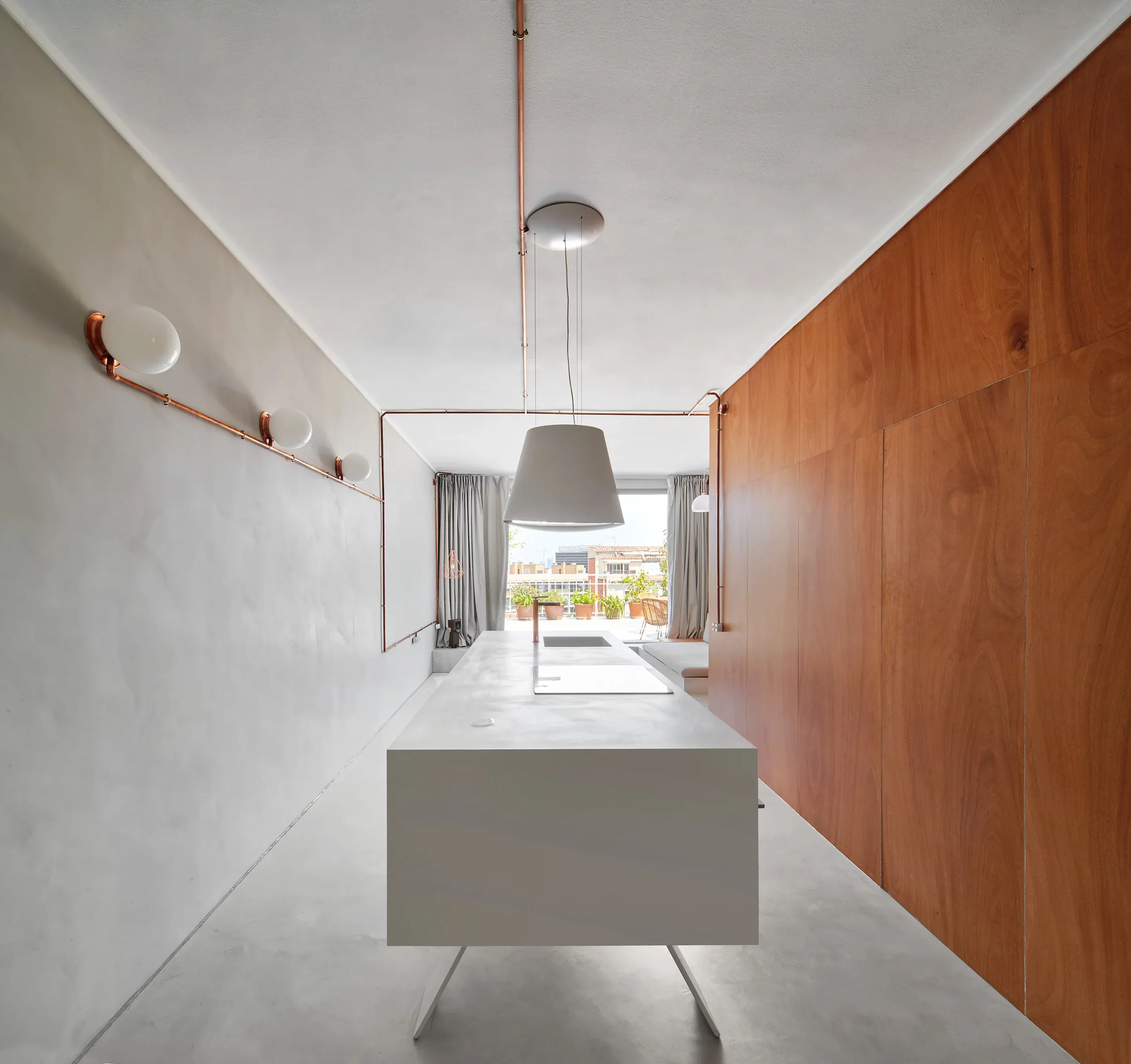 View through Marina Apartment showing white kitchen island, copper lighting, and wooden wall by Cometa Architects in Barcelona