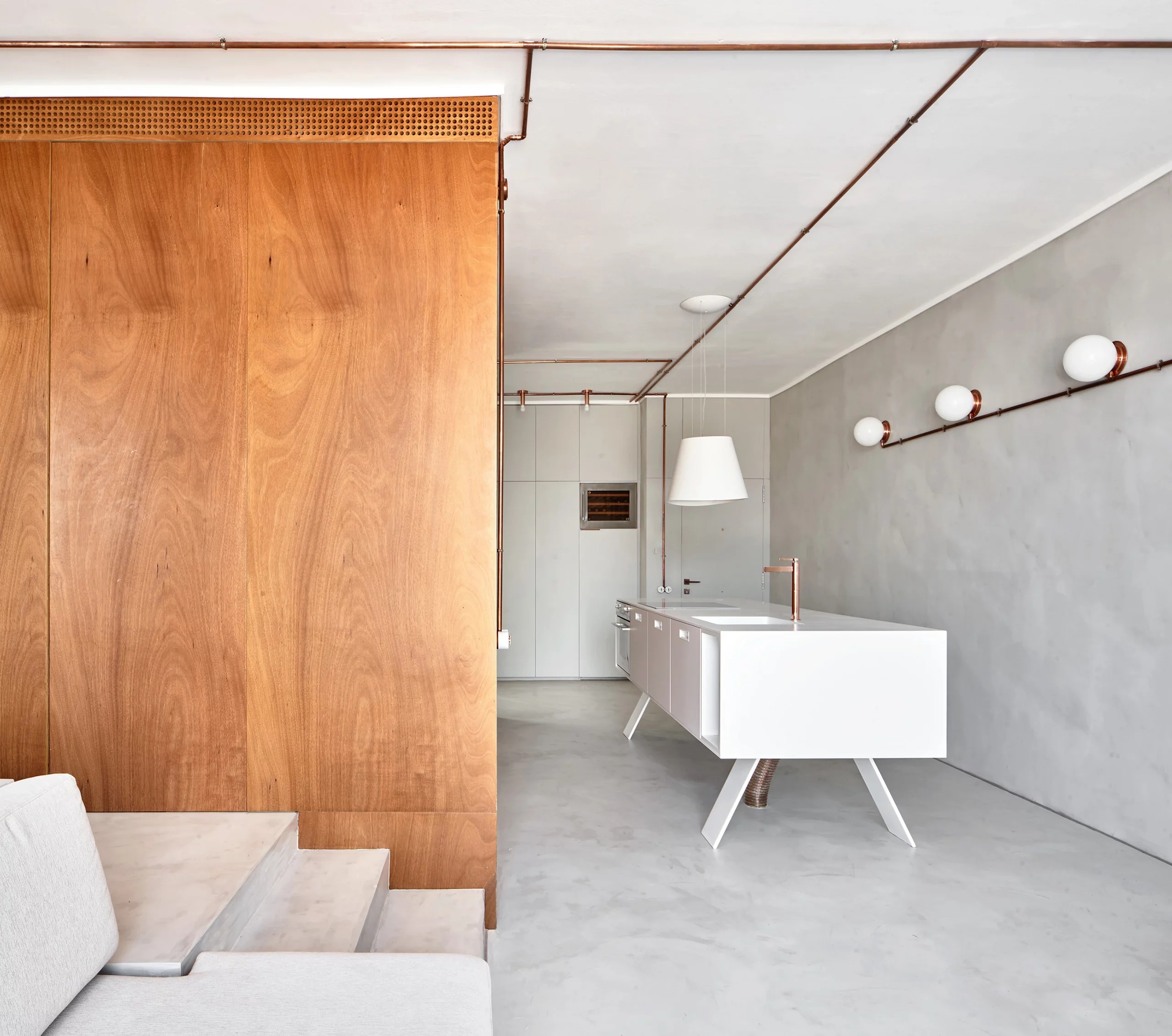 Interior view of Marina Apartment with floating white kitchen island, copper piping, and wooden wall by Cometa Architects in Barcelona