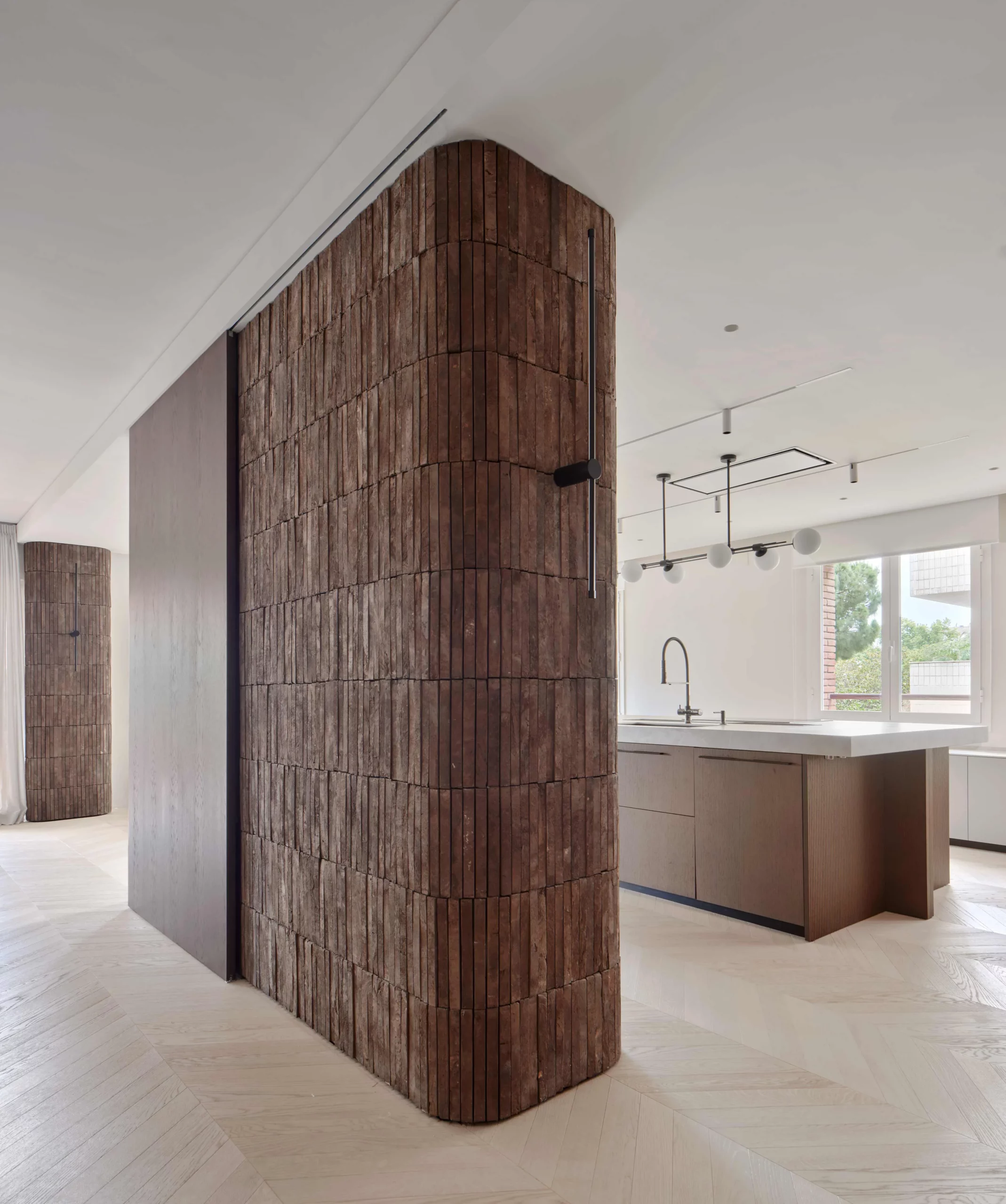 Curved dark brick wall beside modern kitchen island with brushed metal faucet and chevron oak flooring — The Barros Project by Cometa Architects.